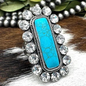 May include: A ring featuring a rectangular turquoise stone with dark veining, set in a silver-toned metal frame. The central stone is surrounded by a border of small, clear, round faceted crystals arranged in a scalloped, oval-shaped pattern.