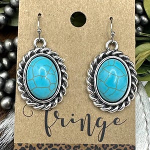 May include: A pair of turquoise-colored oval earrings with a silver-colored rope-like border. The earrings are displayed on a brown card with the word "Fringe" in a handwritten font. The card has a leopard print border.