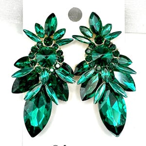 May include: A pair of emerald green statement earrings. Each earring features a cluster of faceted, teardrop-shaped and oval-shaped green crystals, with smaller crystals surrounding a central, larger stone. The earrings are set on a gold-toned metal backing. The text "Fringe after Dark" is visible.
