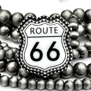 May include: A layered necklace featuring matte silver-colored beads. The focal point is a Route 66 shield-shaped pendant with the words "ROUTE 66" and the number "66" in black on a white background. The pendant has a decorative silver border.