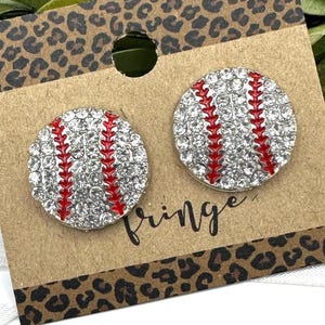 May include: A pair of baseball-themed stud earrings. Each earring is a round, silver-colored stud with a red stitched design, resembling a baseball. The earrings are set against a brown card with the word "Fringe" and a leopard print border.
