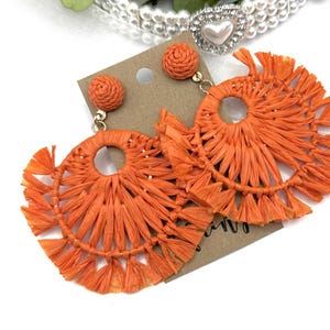 May include: A pair of orange tassel earrings with a woven circle design. The earrings are handmade and have a bohemian style.