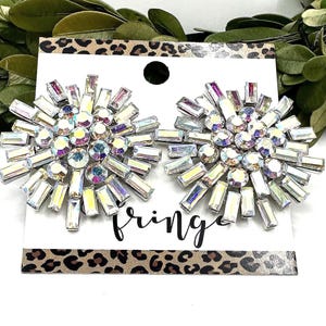 May include: A pair of sparkling, crystal-like earrings with a starburst design. The earrings are set on a white card with the word "fringe" in script. The card has a leopard print border.