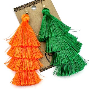 May include: A pair of tiered tassel earrings attached to a brown card. One earring is bright orange and the other is vibrant green. Each earring features four distinct layers of fine thread tassels suspended from a small gold-tone metal hook.