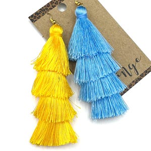 May include: A pair of tiered tassel earrings featuring one bright yellow tassel and one light blue tassel. Each earring is constructed with three distinct layers of fine thread and attached to a gold-toned metal hook. The earrings are displayed on a brown card.