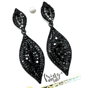 May include: A pair of black, leaf-shaped dangle earrings. Each earring is covered in small, sparkling black rhinestones. The earrings hang from a small, black loop. The earrings are on a white background with a small tag that says "fringe after dark".