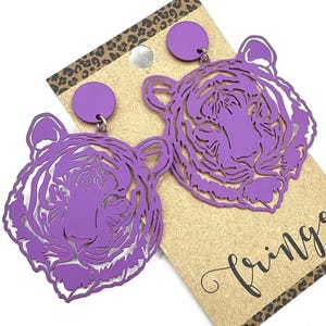 May include: Purple tiger head earrings with a cut-out design. The earrings are attached to a small purple circle at the top and hang from a silver-colored hook. The earrings are displayed on a brown tag with the word "Fringe" in cursive.