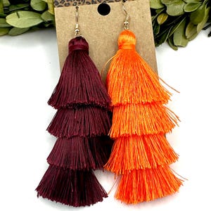 May include: A pair of tiered tassel earrings featuring one deep burgundy tassel and one bright orange tassel. Each earring consists of four stacked layers of fine thread, suspended from a gold-toned metal hook, displayed on a brown card.