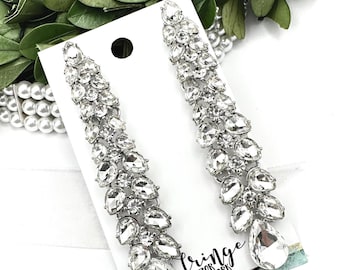 May include: A pair of long, silver dangle earrings with clear crystal teardrop-shaped stones. The earrings are displayed on a white card with the text "fringe after dark".