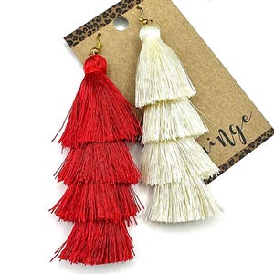 May include: A pair of tiered tassel earrings, one in bright red and one in off-white, attached to a brown cardboard display card with a leopard print border. Each earring features four stacked layers of fine thread fringe and gold-tone hooks.