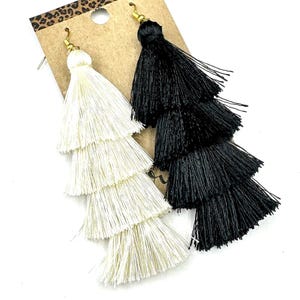 May include: A pair of long, tiered tassel earrings attached to a brown cardboard display card. One earring is white and the other is black, both featuring four distinct layers of fine thread fringe and gold-toned metal earring hooks at the top.