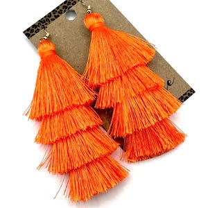 May include: A pair of bright orange tiered tassel earrings featuring four distinct layers of fine thread fringe. Each earring is attached to a gold-toned metal hook and is displayed on a brown cardboard backing card with a leopard print border.