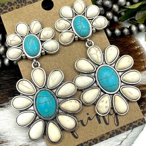 May include: A pair of flower-shaped earrings with a turquoise center stone and off-white petals. The earrings have a silver-colored metal setting and are displayed on a brown card with a leopard print border.