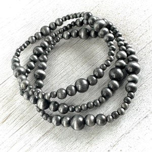 May include: Three silver beaded bracelets stacked on a white wooden surface. The beads are round and have a brushed metal finish.