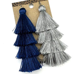 May include: A pair of tiered tassel earrings attached to a brown cardboard display card with a leopard print top border. One earring is deep navy blue and the other is metallic silver, both featuring four stacked layers of fine thread fringe.