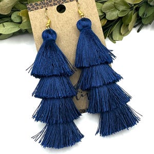 May include: A pair of long, tiered navy blue tassel earrings featuring four stacked layers of fine thread. The earrings are attached to a rectangular brown cardboard display card with a leopard print border at the top and gold-toned hooks.
