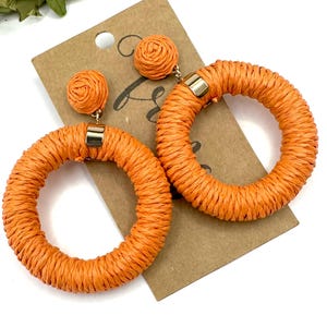 May include: A pair of bright orange hoop earrings featuring a woven raffia texture. Each earring consists of a circular hoop attached to a small, round stud component. A gold-toned metal connector secures the woven material on each hoop.