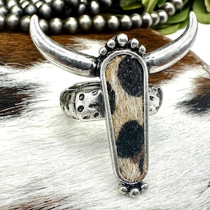 May include: A silver-toned ring featuring a long, rectangular centerpiece with a cowhide pattern and a pair of curved horns. The ring is set against a backdrop of a cowhide pattern and greenery.