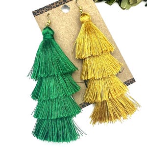 May include: A pair of tiered tassel earrings, one in vibrant green and one in bright yellow, attached to a brown cardboard display card. Each earring features three stacked layers of fine thread fringe suspended from gold-toned metal hooks.