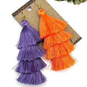 May include: A pair of tiered tassel earrings featuring one purple and one bright orange tassel, each constructed from multiple layers of fine thread. The earrings are attached to gold-toned metal fishhook wires and displayed on a brown card.