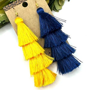 May include: A pair of long, tiered tassel earrings attached to a brown cardboard display card. One earring is bright yellow and the other is deep navy blue. Each earring features four stacked layers of silk-like thread tassels with gold-tone hooks.