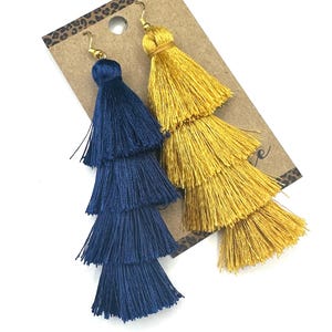 May include: A pair of long, tiered tassel earrings featuring one deep navy blue tassel and one bright golden yellow tassel. Each earring is constructed with four distinct layers of fine thread, suspended from gold-toned metal earring hooks.