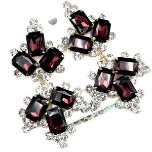 May include: A pair of statement earrings featuring large, rectangular, deep purple faceted crystals surrounded by clusters of small, clear, round rhinestones. The earrings are attached to a display card with the text After Dark printed on it.