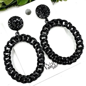 May include: A pair of black hoop earrings. Each earring features a small, round stud with a cluster of black crystals. The hoops are made of linked chain segments, also in black, and are embellished with crystals.