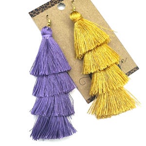 May include: A pair of tiered tassel earrings attached to a brown cardboard display card. One earring is vibrant purple and the other is bright yellow. Each earring features four stacked layers of fine thread tassels and gold-toned metal hooks.