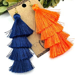May include: A pair of tiered tassel earrings featuring one deep navy blue and one bright orange tassel, each constructed with four stacked layers of fine thread. The earrings are attached to a brown cardboard display card with a leopard print top.