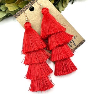 May include: A pair of vibrant red tiered tassel earrings featuring four distinct layers of fine thread. The earrings are attached to a rectangular brown card with a leopard print border at the top and a partial black script logo on the side.