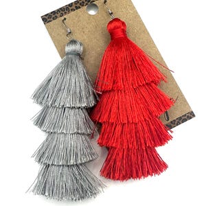 May include: A pair of tiered tassel earrings attached to a brown display card. One earring is silver and the other is bright red. Each earring features four distinct layers of fine, silky thread tassels suspended from a silver-toned metal hook.