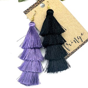 May include: A pair of tiered tassel earrings featuring one purple and one black earring, each with four layers of silky thread. The earrings hang from gold-toned metal hooks and are attached to a brown cardboard display card with the word fringe printed.