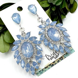 May include: A pair of silver dangle earrings with large, light blue, teardrop-shaped crystals. The crystals are surrounded by smaller, clear crystals.