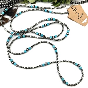 May include: A long necklace featuring small, silver-colored beads interspersed with turquoise-colored beads. The necklace is displayed on a white surface with a brown tag that reads "Living". The necklace is a western-style accessory.