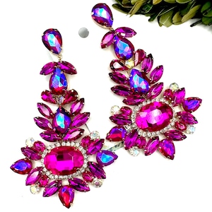 May include: A pair of long, chandelier-style earrings featuring vibrant fuchsia and iridescent purple faceted crystals. The design includes a large central oval stone surrounded by smaller marquise and pear-shaped crystals set in metal.