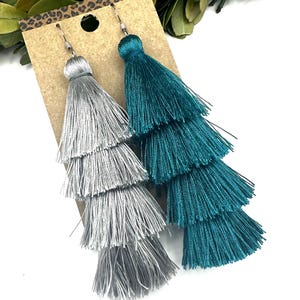 May include: A pair of tiered tassel earrings featuring one silver and one teal earring. Each earring consists of four stacked layers of fine thread fringe attached to a silver-toned metal hook, displayed on a rectangular brown card.