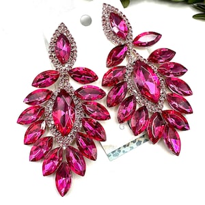 May include: A pair of dazzling fuchsia earrings. Each earring features a central, large, oval-shaped gem surrounded by smaller, leaf-shaped gems. The gems are set in a silver-toned metal, creating a glamorous and eye-catching accessory.