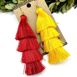 May include: A pair of tiered tassel earrings featuring one bright red earring and one vibrant yellow earring. Each earring is constructed from multiple layers of fine silk-like thread and hangs from a gold-toned metal hook on a brown display card.