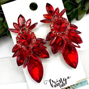 May include: A pair of red crystal earrings with a floral design. The earrings feature large, faceted red crystals arranged in a layered, petal-like pattern. The center of each earring has a cluster of smaller crystals. The earrings are on a white card with the text "fringe after dark".