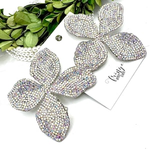 May include: Two large, sparkling flower-shaped earrings. Each earring is covered in iridescent rhinestones, creating a shimmering effect. The earrings are on a white card with the text "Fringe after dark".