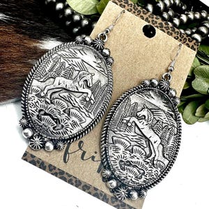 May include: Silver-toned oval earrings with a detailed relief of a horse in a landscape. The earrings have a beaded border and a decorative floral element at the top and bottom. The word "fri" is visible on the tag.