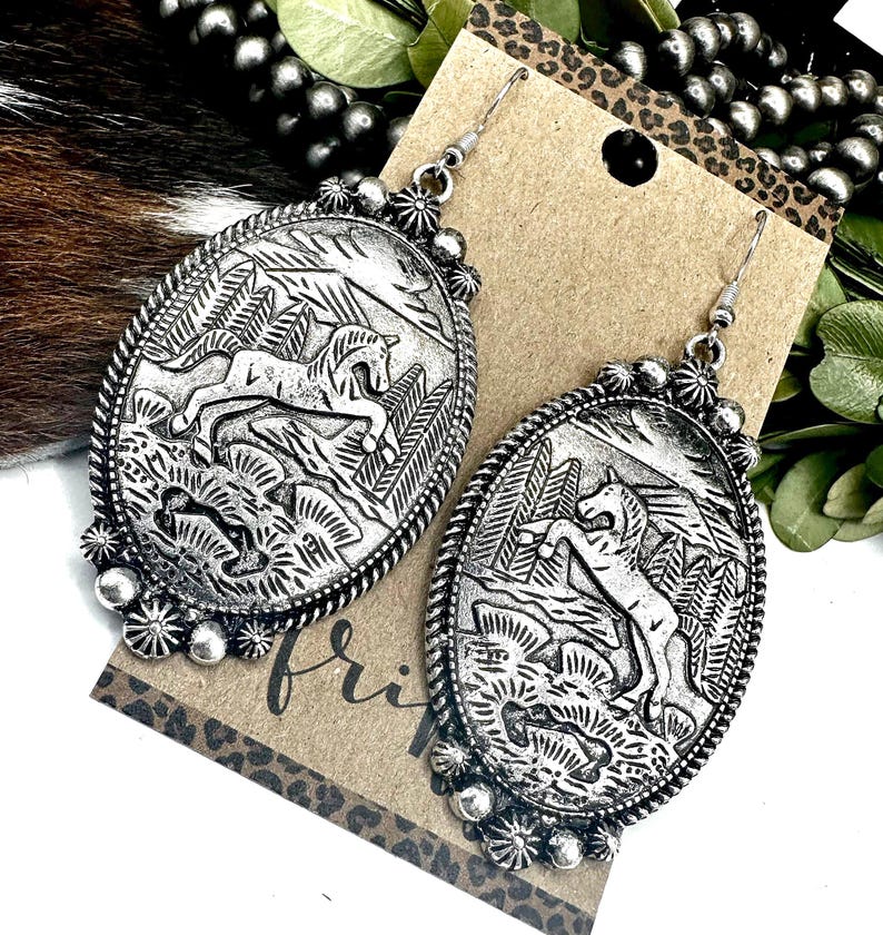 May include: Silver-toned oval earrings with a detailed relief of a horse in a landscape. The earrings have a beaded border and a decorative floral element at the top and bottom. The word "fri" is visible on the tag.