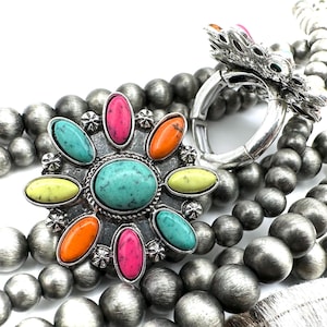 May include: A multi-strand necklace with silver beads and a large silver charm featuring a turquoise stone surrounded by smaller stones in various colors, including pink, orange, yellow, and green.