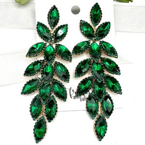 May include: A pair of dangling earrings featuring a leaf design. Each earring is adorned with numerous emerald green, faceted crystals set in a gold-tone metal. The earrings have a post back closure and are displayed on a white card.