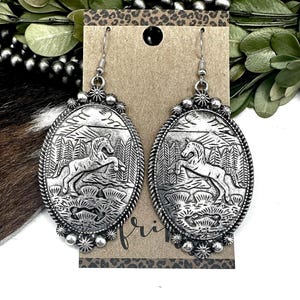May include: Silver-toned oval earrings with a detailed relief of a horse rearing in a forest scene. The earrings have a decorative border and dangle from a simple hook. The background features a brown card with a leopard print.