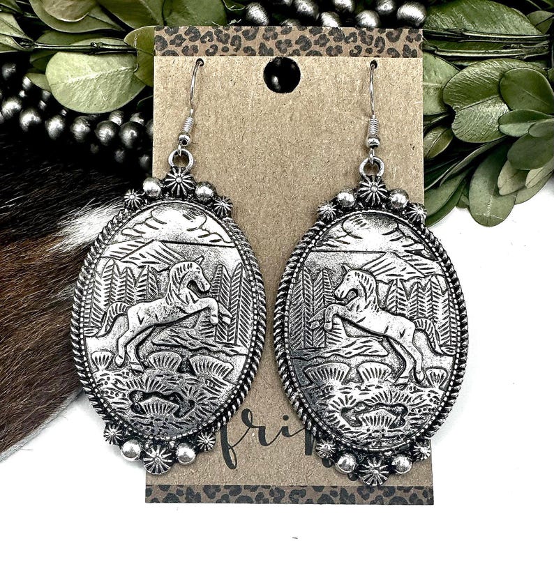 May include: Silver-toned oval earrings with a detailed relief of a horse rearing in a forest scene. The earrings have a decorative border and dangle from a simple hook. The background features a brown card with a leopard print.