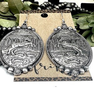 May include: Silver-toned oval earrings with a detailed relief of a horse leaping in a forest scene. The earrings have a beaded border and a decorative top. The design is on a brown card with the word "fri" visible.