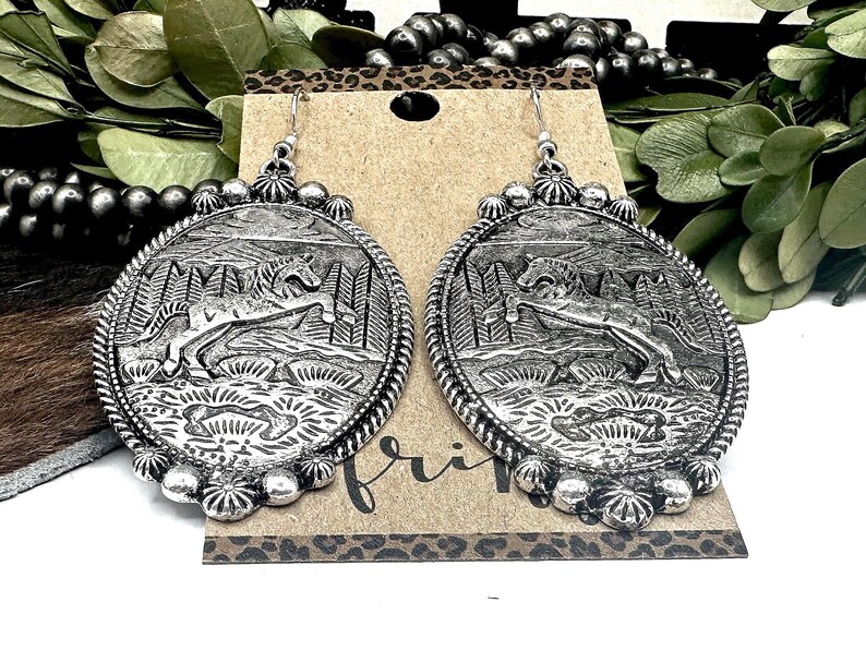May include: Silver-toned oval earrings with a detailed relief of a horse leaping in a forest scene. The earrings have a beaded border and a decorative top. The design is on a brown card with the word "fri" visible.
