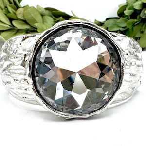 May include: Silver-toned cuff bracelet featuring a large, faceted, clear crystal centerpiece. The bracelet has a textured, hammered metal design around the crystal and on the band. The background includes greenery.